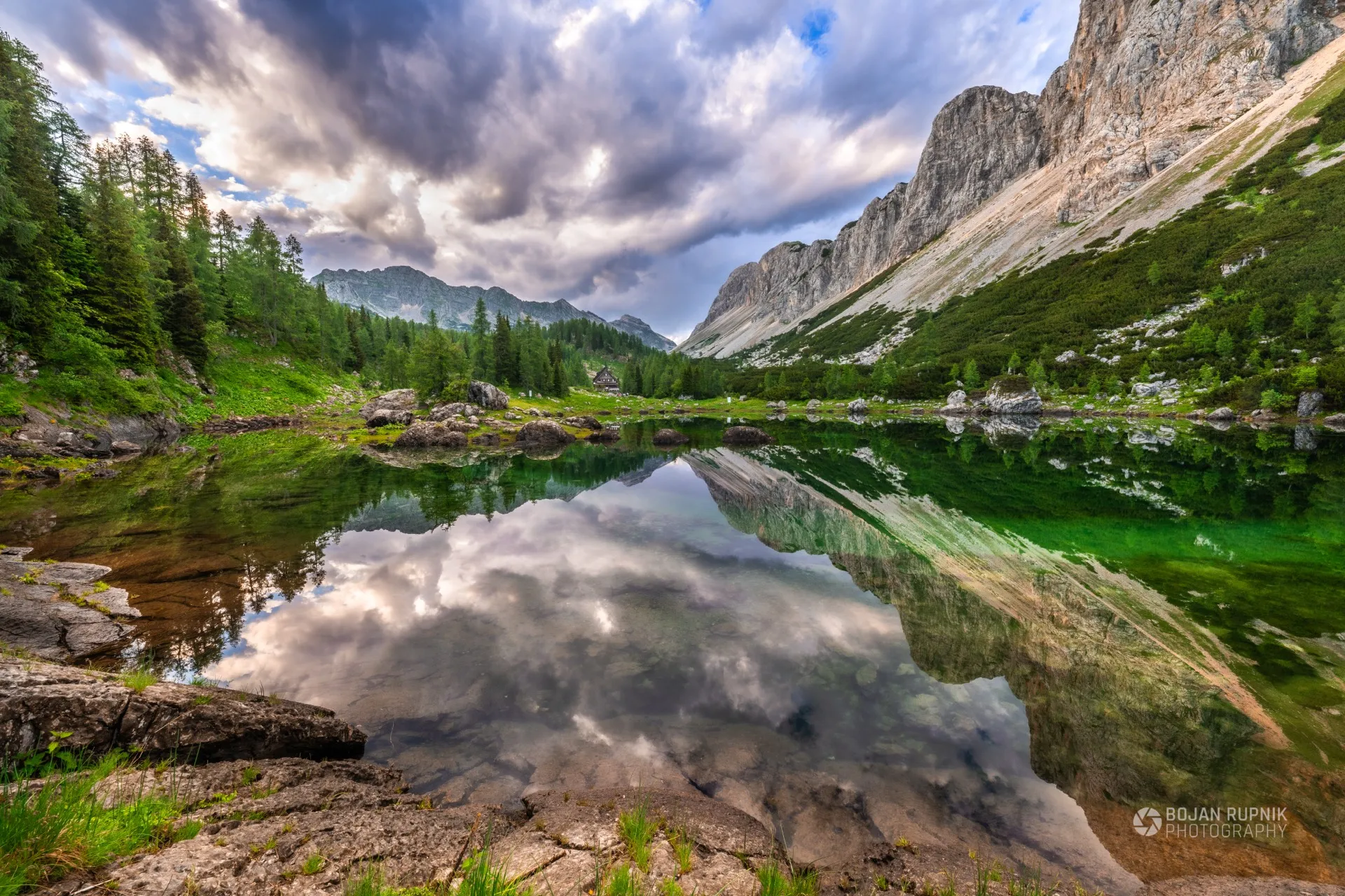 Gorsko jezero z odsevom oblakov in skalnatih sten med macesni