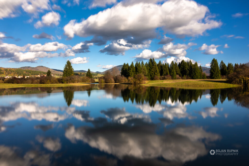 Simetrija odseva na vodni gladini poplavljenega Obrha v Loški dolini z krasnimi belimi oblaki